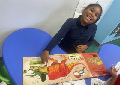 A girl smiles as she points at an open book with a diagram of a Triceratops.