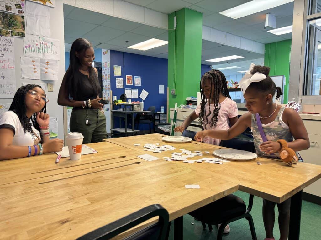 Jakira Swift, smiling, stands above three students in her program as they play a game spread out on the table in front of them.